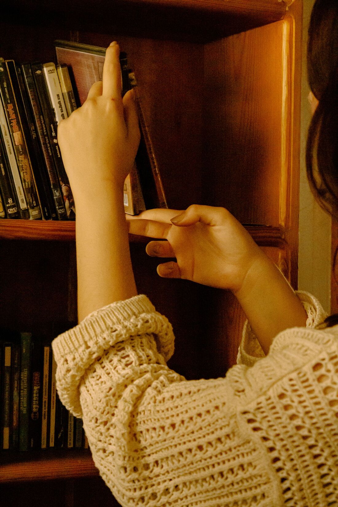 A woman in a cream sweater reaching for a book on a wooden shelf