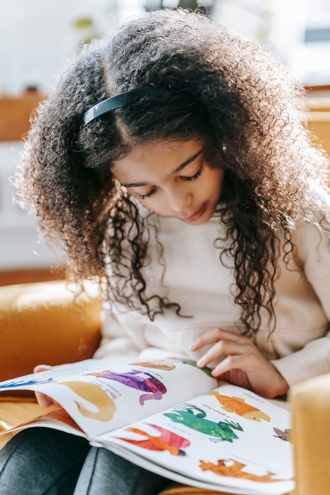 A girl with curly hair reading a colorful picture book in an armchair