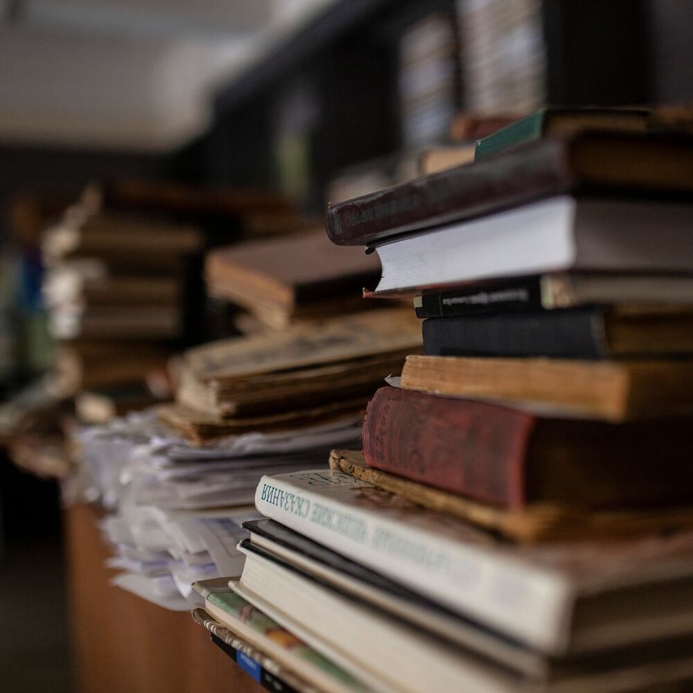 A leaning pile of old hardcover books beside a bookcase